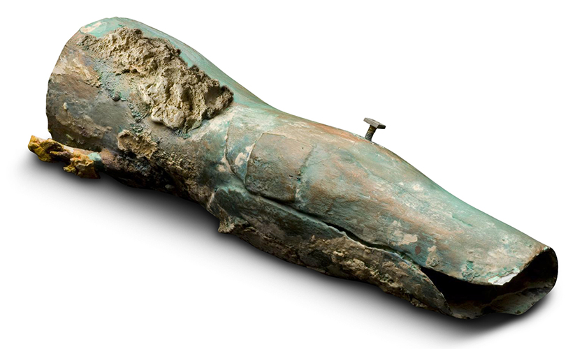 A tubular object of greenish, encrusted metal, shaped like a person’s lower thigh and upper calf. The head of a nail or screw juts from the surface.