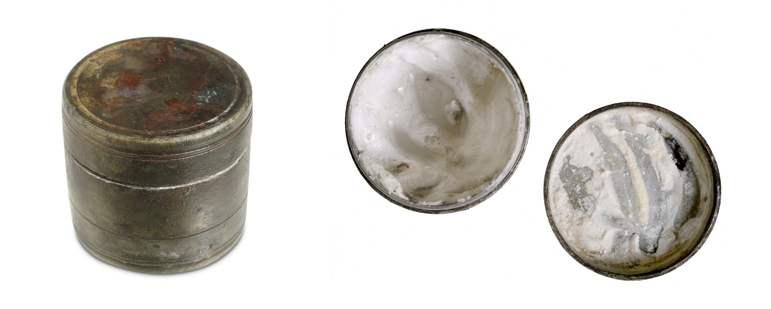 A darkened metallic container with a cylindrical shape. The container’s closed lid has splotches of a coppery red. 