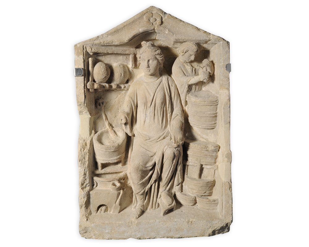 A bas relief featuring a female figure sitting inside an architectural space. Around her are various baskets and cauldrons on shelves. Another figure is visible working in the background.