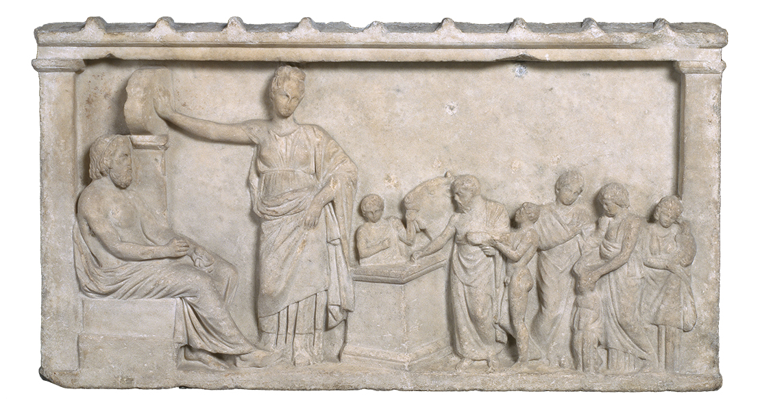 A horizontal bas relief of two tall figures, a seated male and a standing female, and several smaller figures grouped to the right. All figures wear togas.