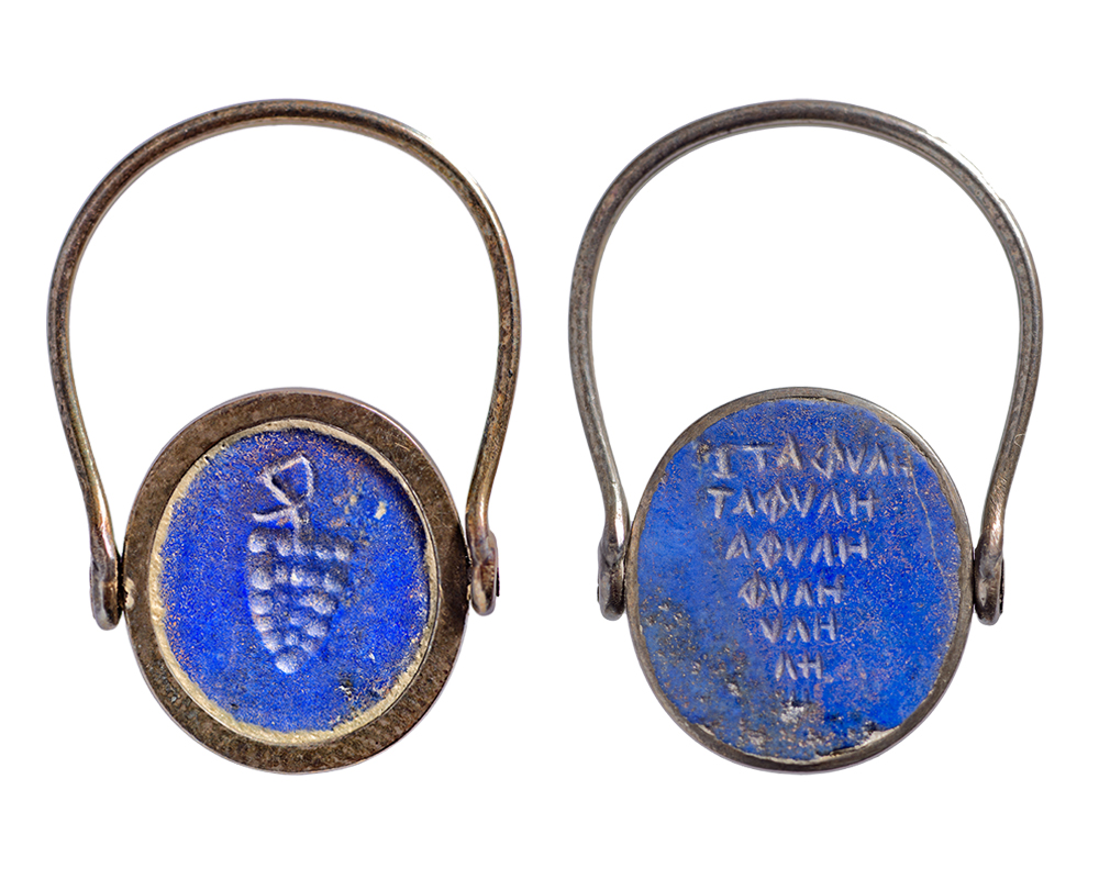 The front and back of a gold ring inset with a blue circular stone. On the left, the circle is inscribed with a bunch of grapes; the right shows seven lines of Greek text in the shape of a downward pointing triangle.