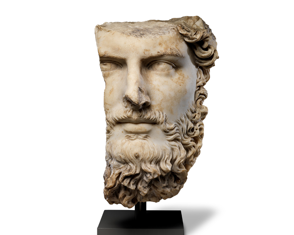 White stone head of a man with orange discoloration. The left side of the face, including the ear, half of the eye, and the crown of the head are missing. A curly moustache frames the lips while a beard covers his neck.