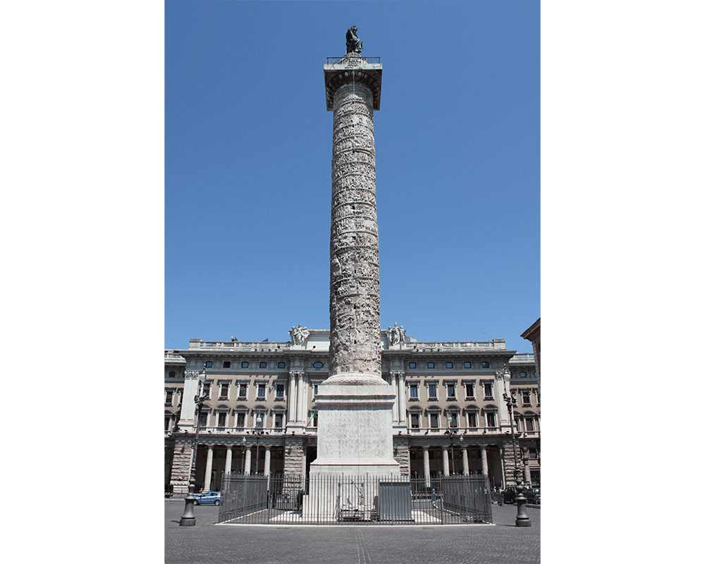 An imposing column decorated with carved figures along a winding spiral. A greenish statue stands atop the column, which has been placed outdoors before a large building.