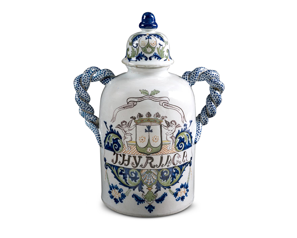 White cylindrical jar with twisted blue handles on either side. The body of the jar is marked “THYRIACA” and decorated with heraldry, putti, and floral motifs. The lid has a design of blue and green foliage, capped by a dark blue finial.