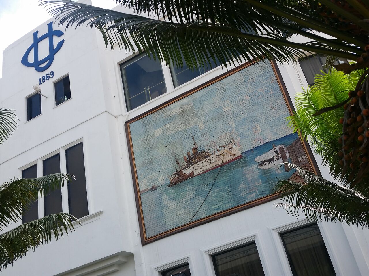 White building with mosaic of ship on ocean and palm tree branches.