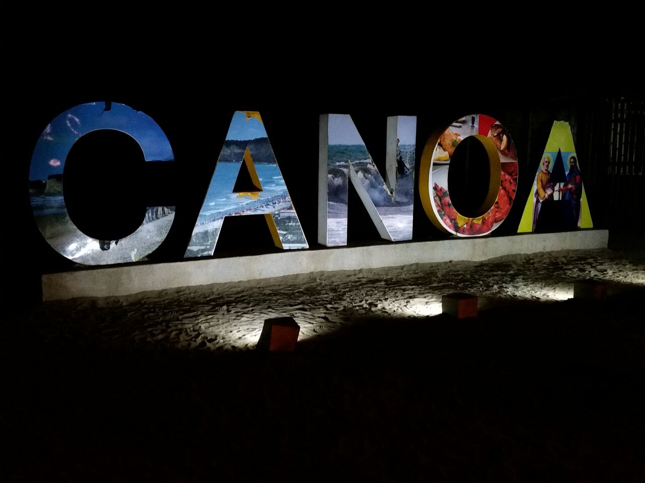 Letters spelling Canoa lit up on the sand at night.