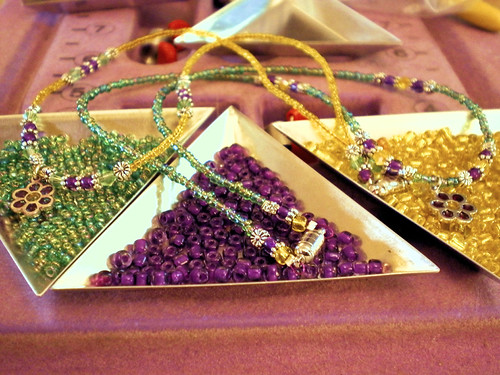 Green, purple, and yellow beads in triangle dishes with necklaces of those colors draped over them holding flower charms.