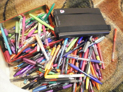 A pile of pens of various colors with a moleskin journal tossed on top