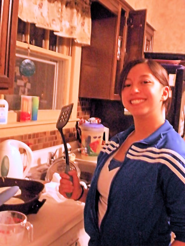 She smiles holding up a spatula while tending an electric skillet of  pancakes