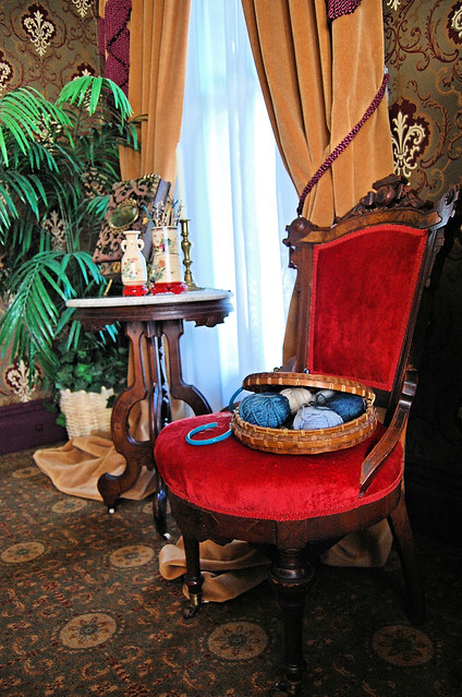 A red velvet chair with a knitting basket full of yarn balls sits next to a marble table. 