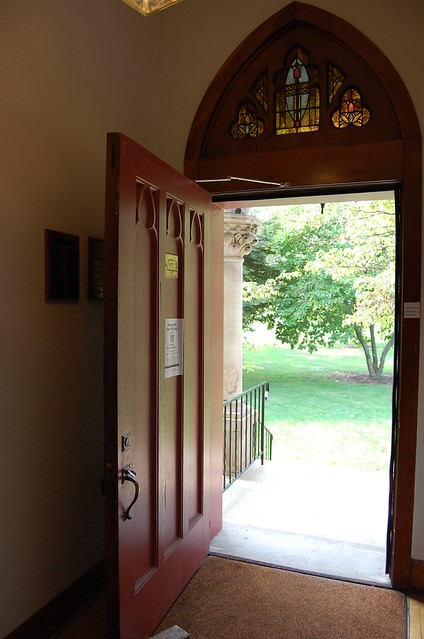A red door stands open to the well manicured lawn. Above the door is a stained glass window.