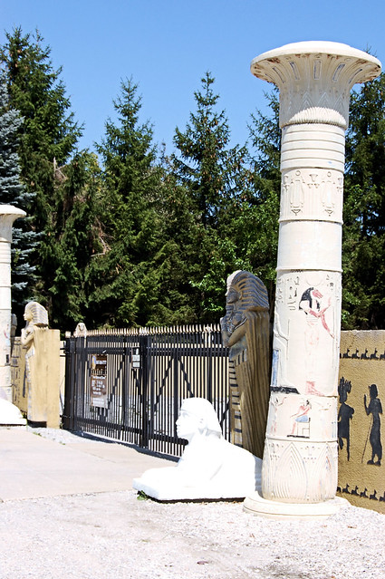 Gate is black wrought iron with golden spear head top, flanking the gate are lotus bloom topped columns and a stark white sphinx