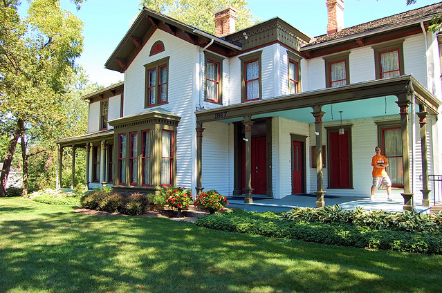 Victorian house in beige trimmed in red & olive with porch ceilings sky blue.  Mike taking a giant step on the main front porch.