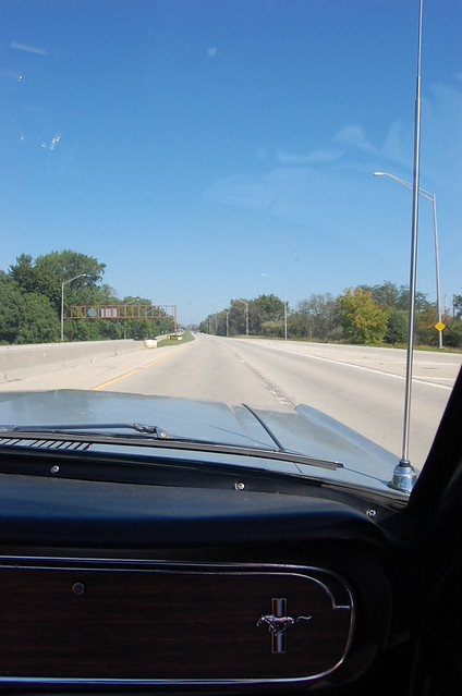 Looking through the windshield to the road with the logo of the classic Mustang horse on the tri-color bar showing in the foreground on the dash.