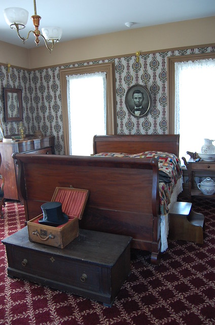 a walnut sleigh style bed with a handmade quilt over it and an oval framed print of Lincoln hanging above the head board at the foot is a travel trunk with a black tall hat