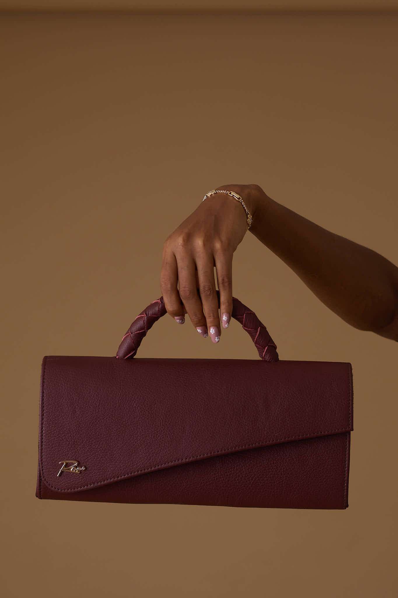 Close-up of a burgundy leather handbag being held by a hand with a gold bracelet against a solid brown background.