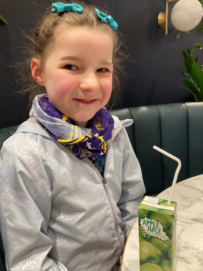 M with a cheeky smile. She is wearing a running jacket and buff and has jade green bows in her hair. She is sitting at a table with a carton of apple juice.