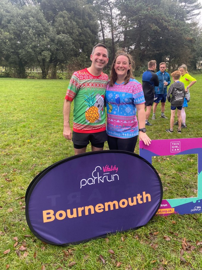 Tamsyn and Stuart standing behind a sign that says Bournemouth parkrun. Stuart has on a red and green Christmas t-shirt. Tamsyn is wearing a pink and blue festive t-shirt with budgies on it.