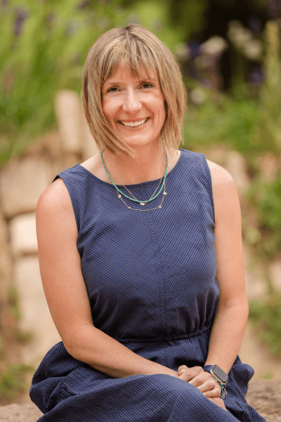 A smiling Nancy sitting outdoors on a stone surface. she has light skin and chin-length light brown hair with bangs that angle slightly across her forehead. she is wearing a sleeveless, ankle length dark blue dress with very fine vertical stripes and a gather waist. her hands are resting losely together in her lap.