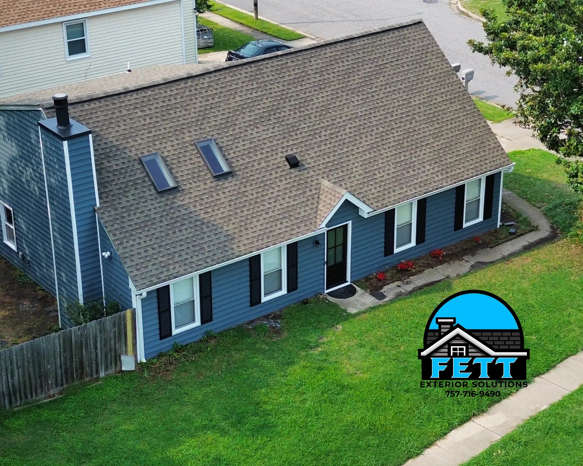 Aerial view of a blue two story house with a brown shingled roof dormer skylights and a front lawn logo overlay in the bottom right corner