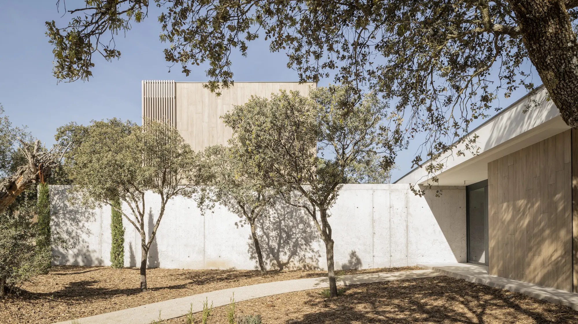 Fachada de vivienda contemporánea de FH2L Arquitectos en Villanueva de la Cañada, Madrid, integrada en un jardín con árboles, hormigón visto y revestimiento de madera.
