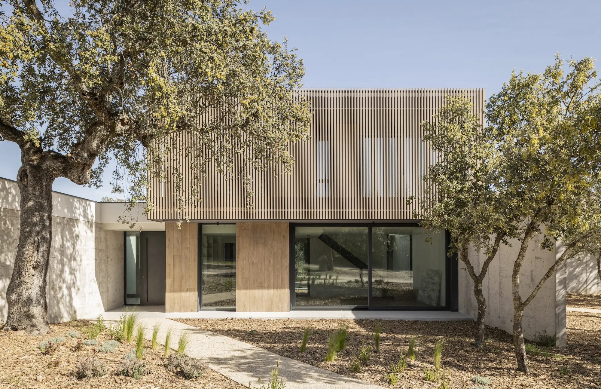 Fachada principal de CASA TG3, vivienda unifamiliar contemporánea de FH2L Arquitectos en Villanueva de la Cañada, Madrid, con celosía vertical, madera y hormigón visto.