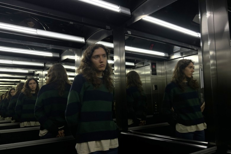 A young woman with curly hair stands alone in an elevator, looking serious. The reflective walls create multiple reflections, enhancing the mood of the scene.
