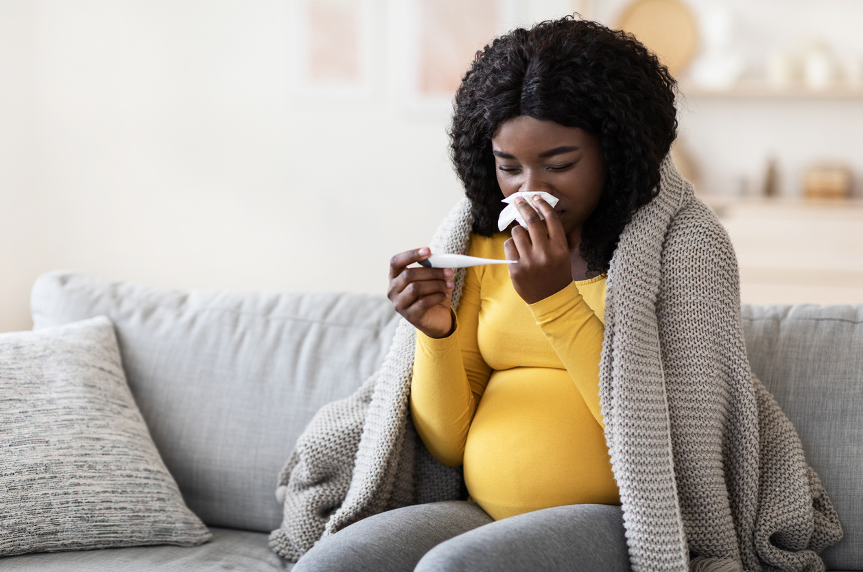 A pregnant individual wearing a blanket around her shoulders while holding a tissue to their nose in one hand and a thermometer in the other on a couch -- Une personne enceinte portant une couverture sur ses épaules, tenant un mouchoir contre son nez dans une main et un thermomètre dans l'autre, assise sur un canapé