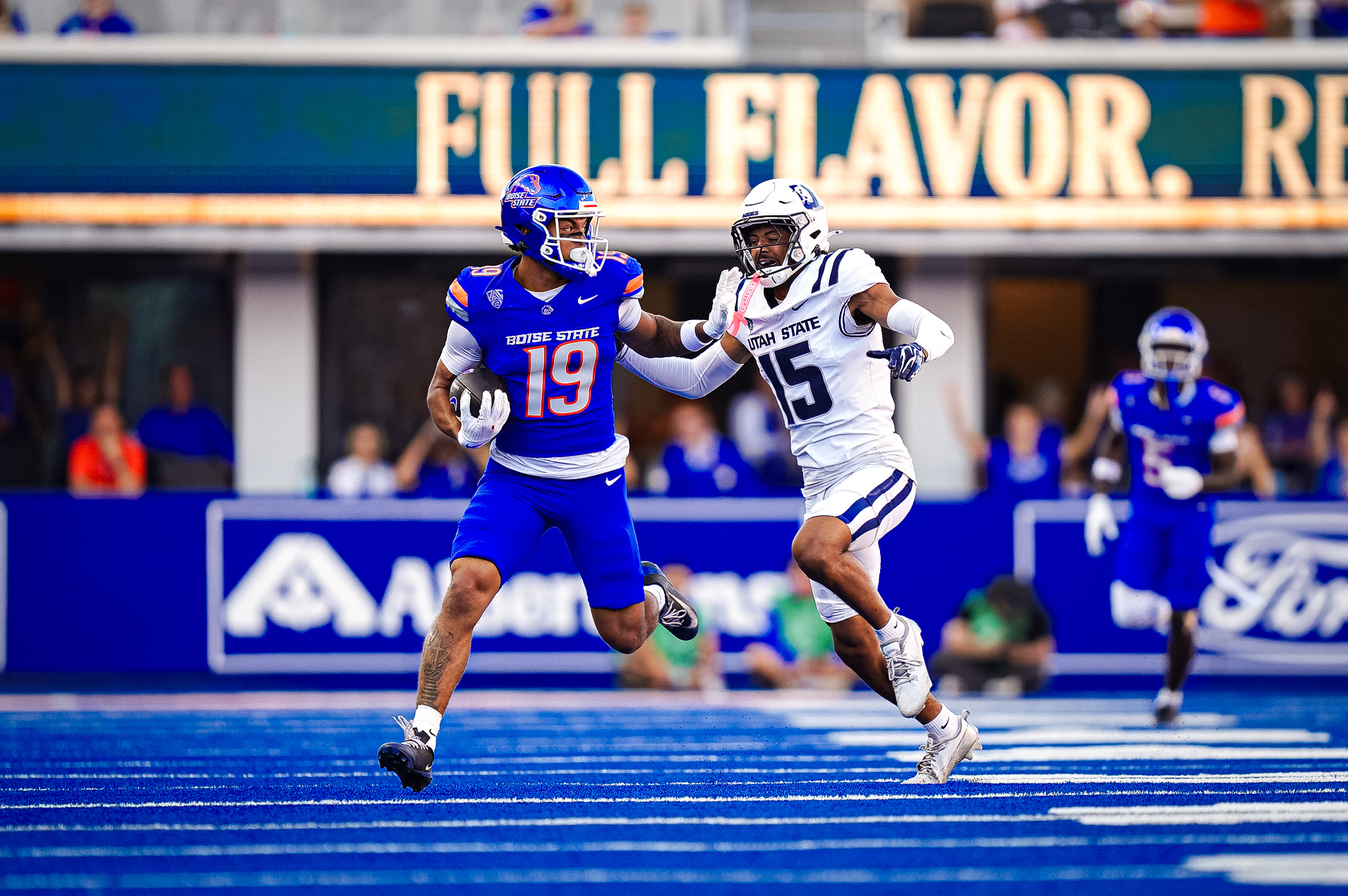 Two football players from opposing teams sprint across a blue field, with one in a blue uniform chasing the other in a white uniform.