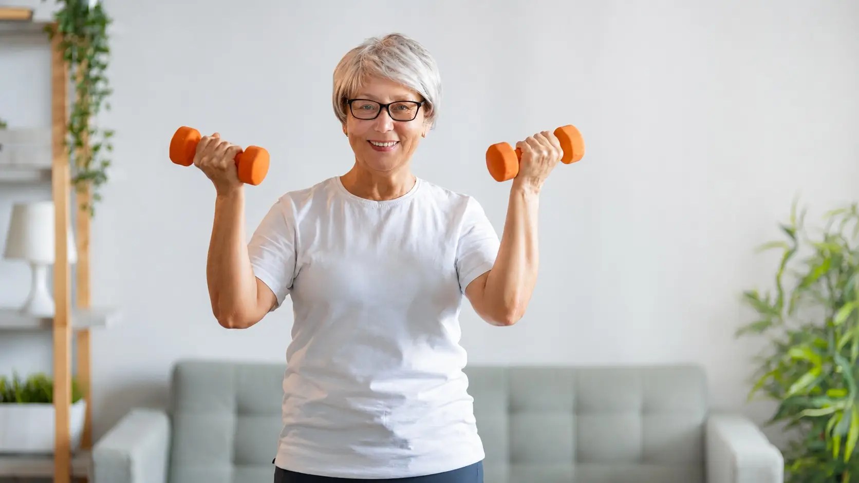 Smiling older woman with glasses lifts orange dumbbells at home for strength training in a bright living room.