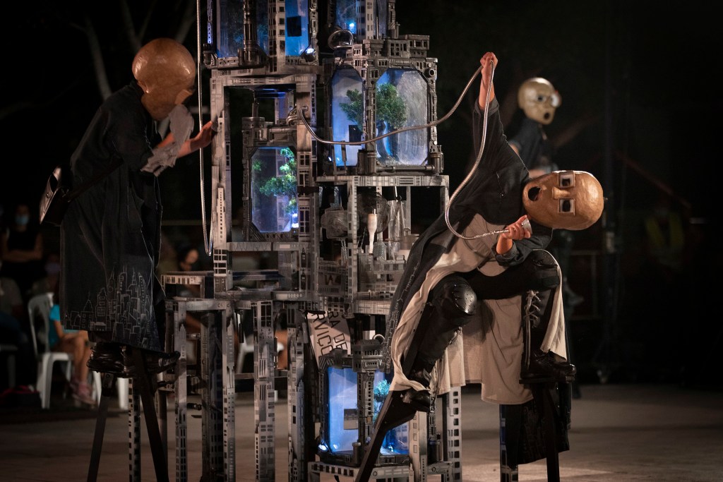 Two performers wearing copper masks manipulate a tall, intricate metal sculpture with blue-lit glass chambers during a nighttime performance.