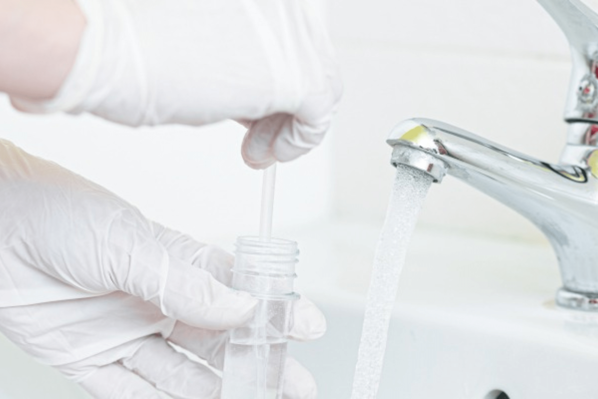 A gloved hand is using a dropper to collect water from a faucet into a clear container for testing.