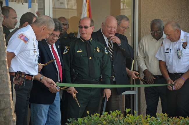 Sheriff Jim Manfre, in uniform, flanked by Palm Coast Mayor Jon Netts and Commissioners Bill McGuire and Bill Lewis to his left, and County Commissioner Nate McLaughlin to his right, at the opening of the Palm Coast precinct at City Market Place last May. The relationship between the city and the sheriff is continuing for the 14th year, and for the foreseeable future. (© FlaglerLive)