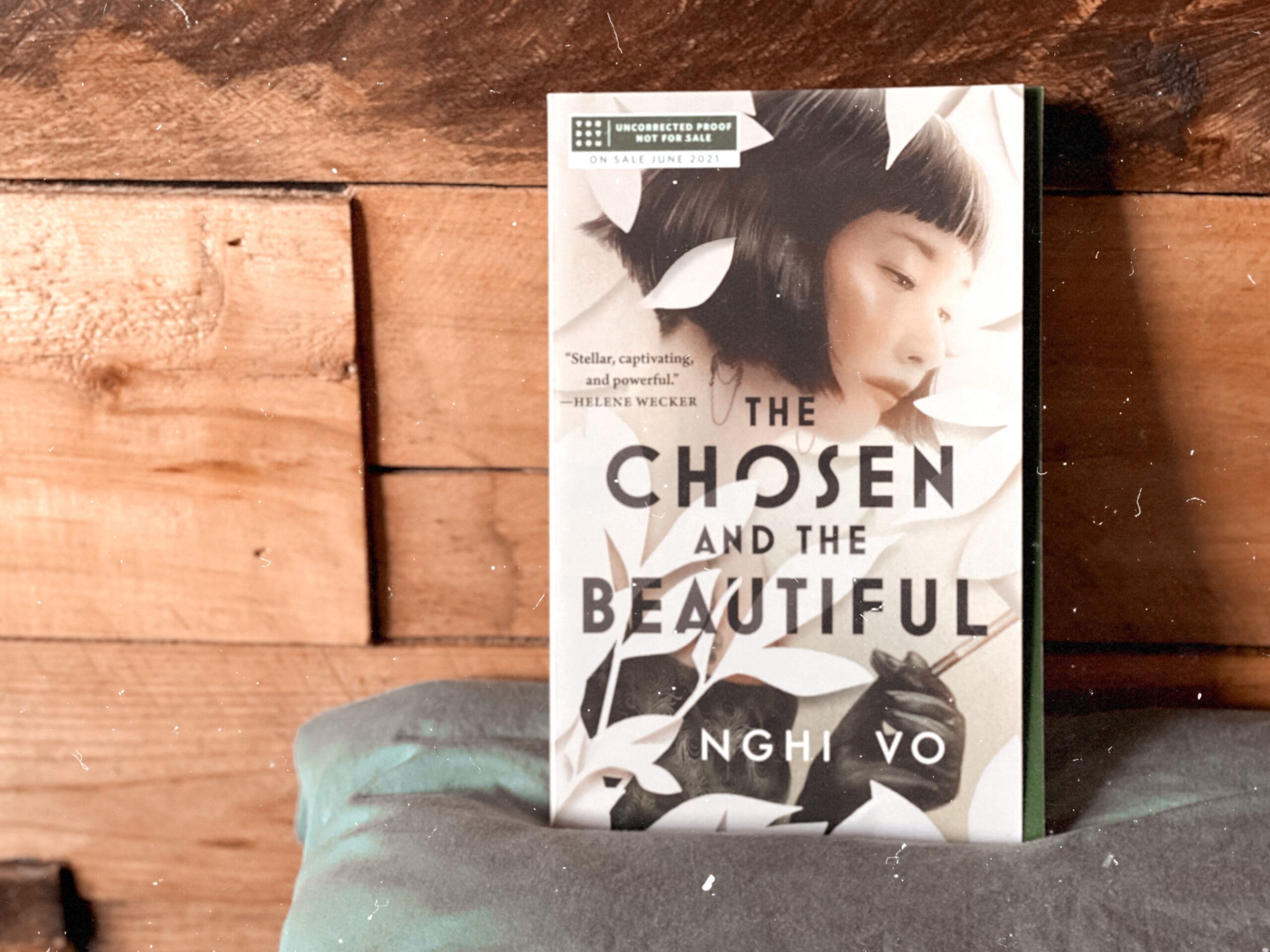 A photograph of an advanced reader copy of The Chosen and the Beautiful by Nghi Vo propped on a pillow and leaning against a reclaimed wood headboard