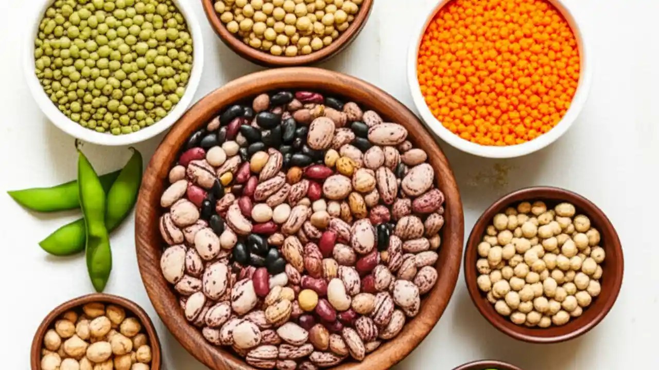 A top-down view of several bowls on a wooden table, each containing a different type of legume, including beans, lentils, peas, and peanuts.