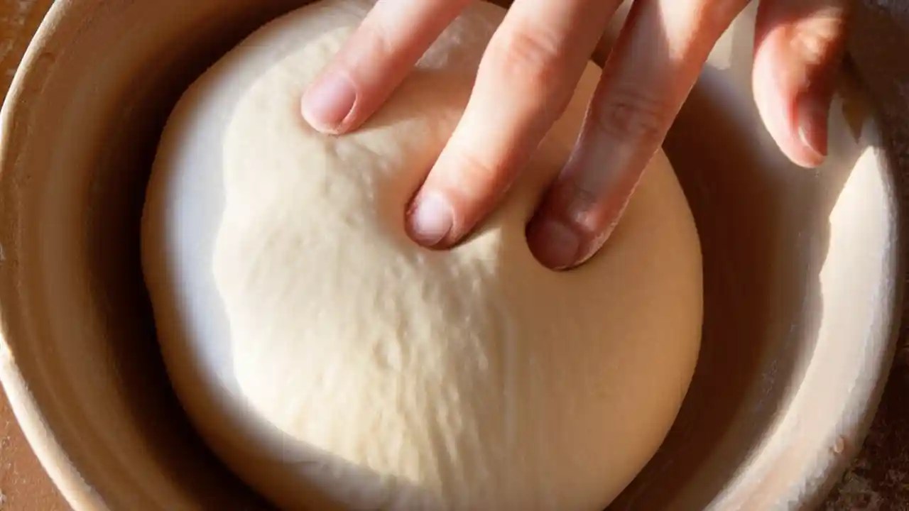 A close-up view of a finger performing the poke test on a smooth ball of risen bread dough in a bowl to check if it's ready for baking.