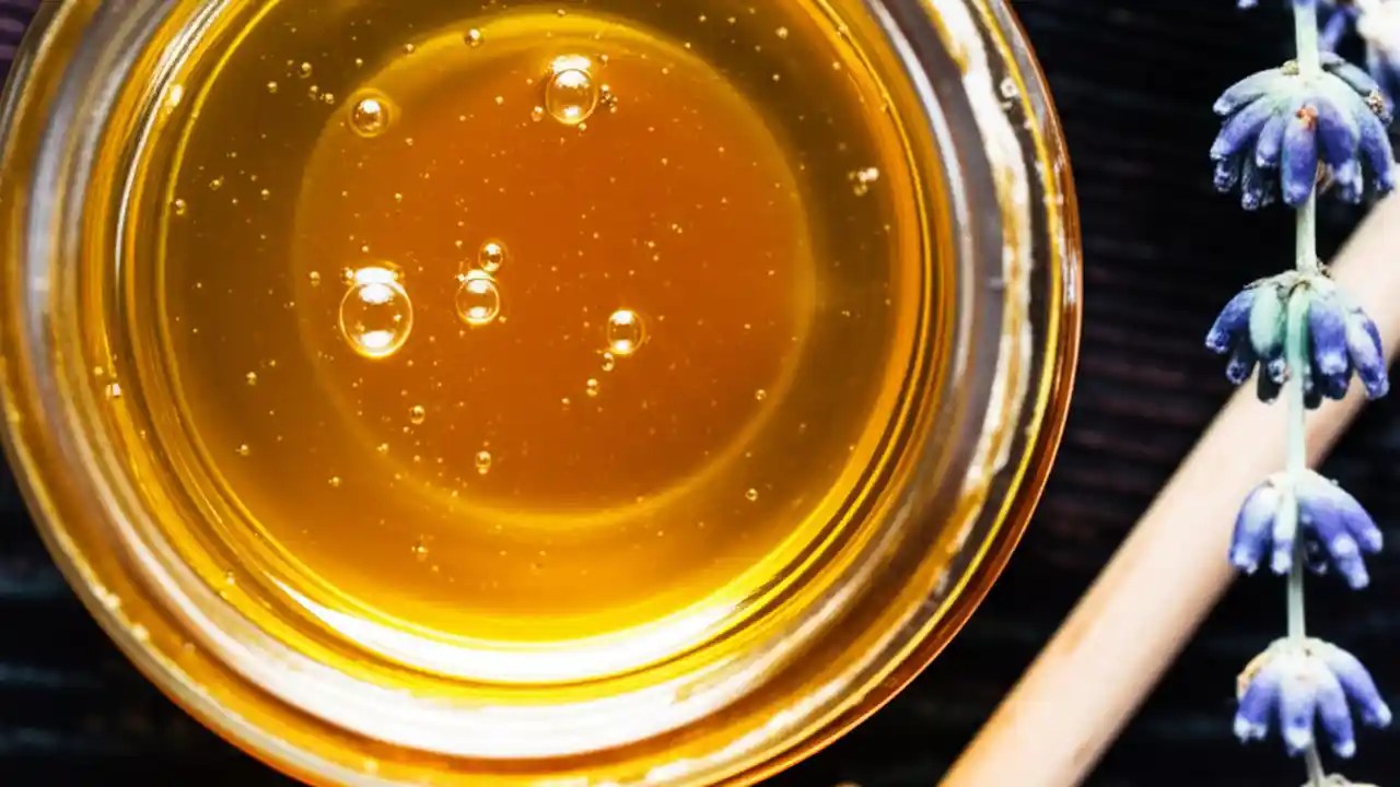 A clear glass jar of golden fermented honey with visible bubbles, sitting next to a honey dipper on a rustic wooden surface.