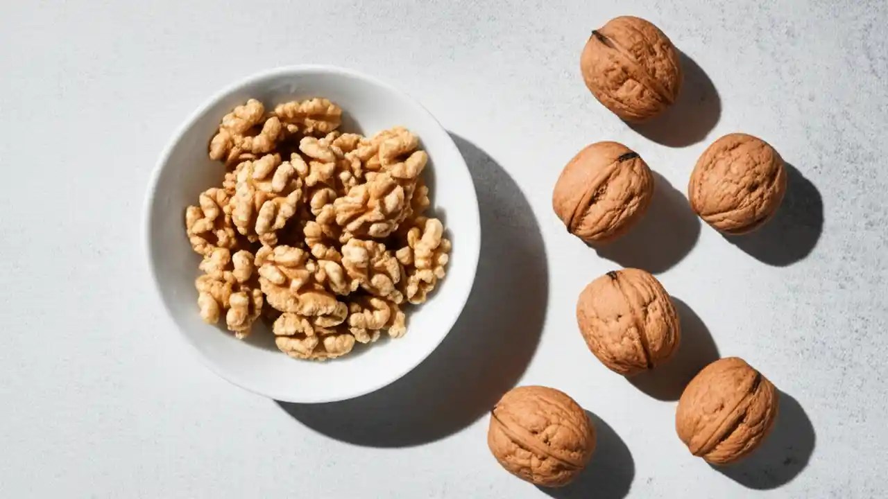 A top-down view of a white bowl containing 14 walnut halves, representing one serving, with 7 whole walnuts next to it on a light surface.