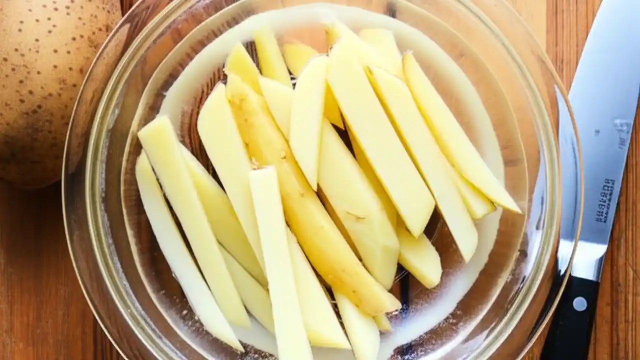 A clear bowl of water with cut french fries soaking, showing the white starch sediment at the bottom, a key step for making crispy fries.