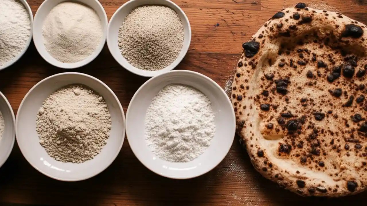 Four bowls showing the different textures of Italian flour types 00, 0, 1, and 2, next to a finished Neapolitan pizza on a wooden table.