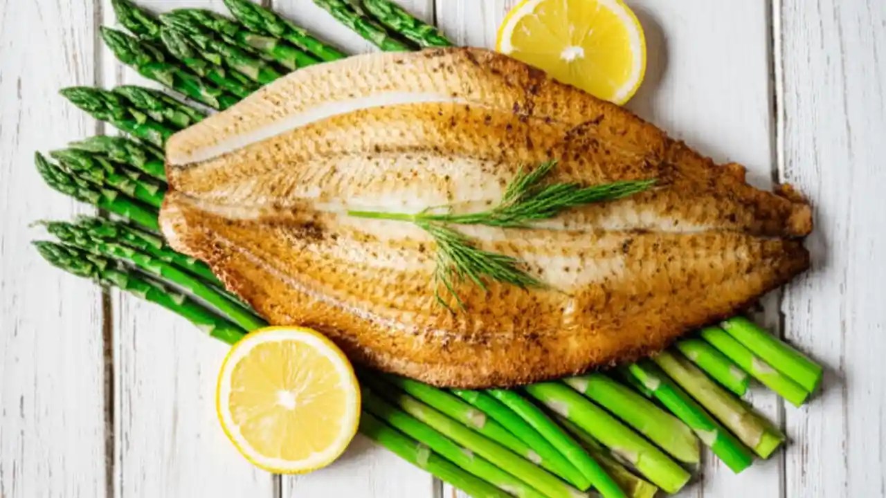 A cooked John Dory fillet, skin-on and golden brown, sits on a white plate next to a lemon wedge, ready to be eaten.