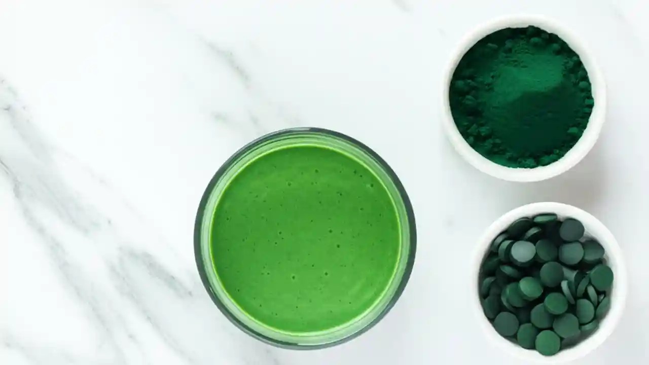 A glass of green spirulina smoothie next to a bowl of spirulina powder and a bowl of tablets, illustrating different daily dosage forms.