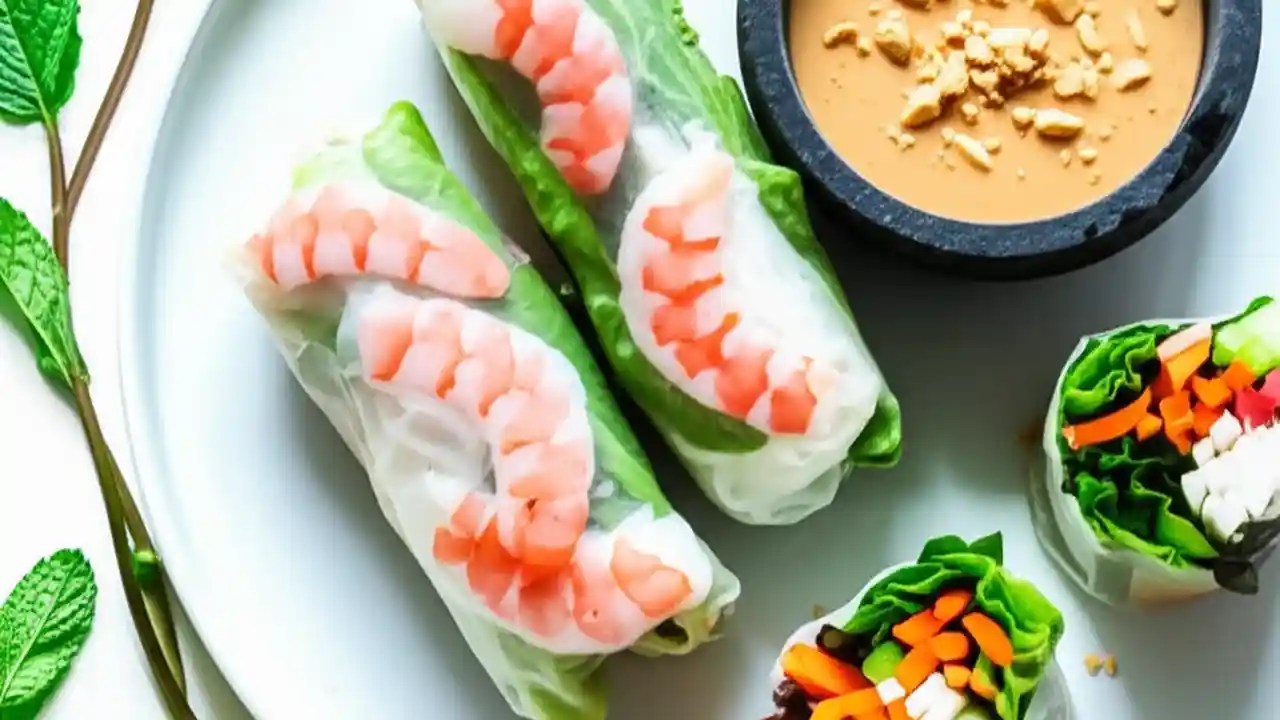 A platter of fresh Thai salad rolls, one sliced open to show the shrimp and vegetable filling, served alongside a bowl of creamy peanut sauce.