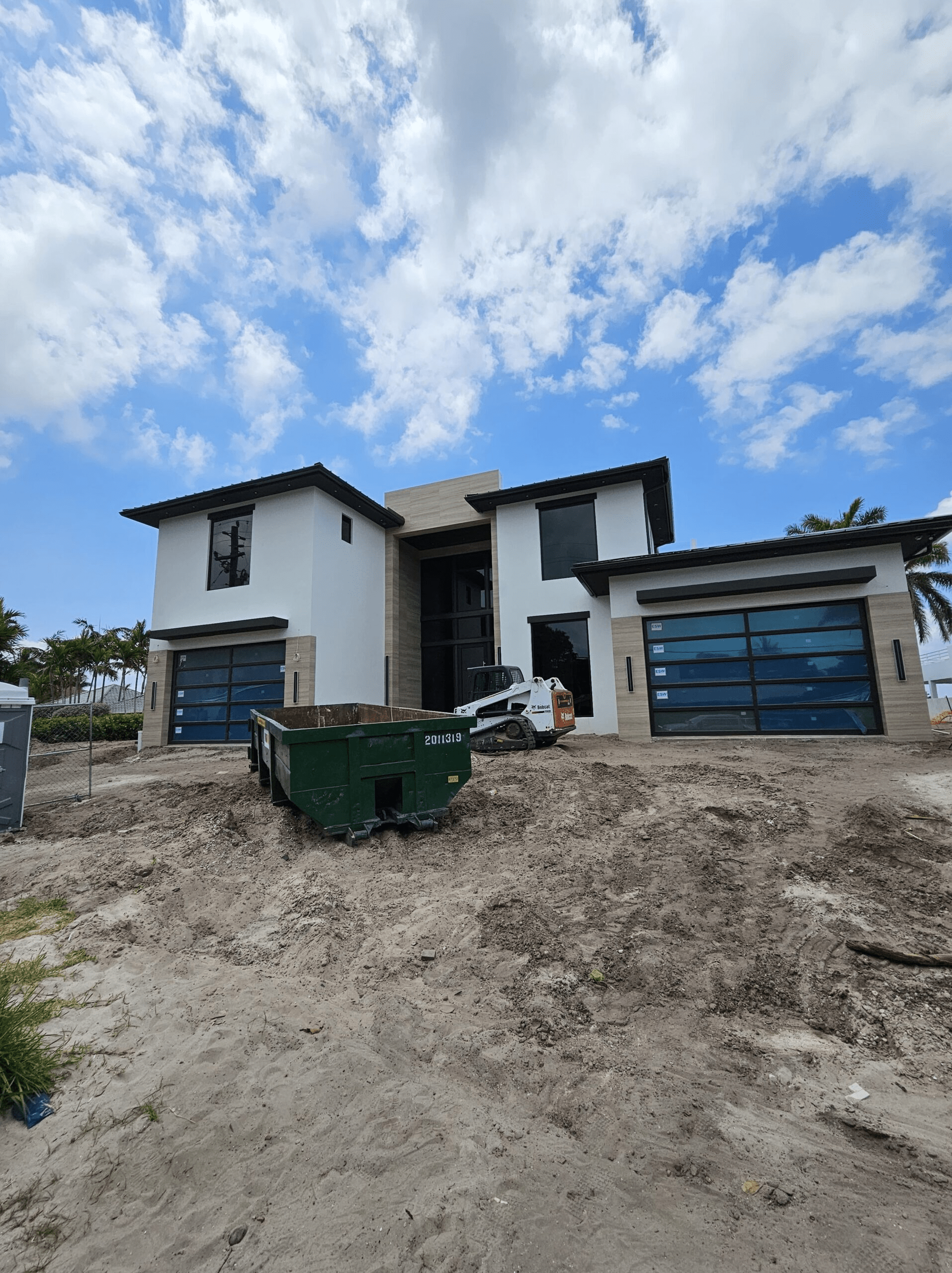 Miami construction site. Dirt and construction supplies sit in the foreground and a house under construction is in the background. The house is white with dark brown trim. The sky is partially cloudy.