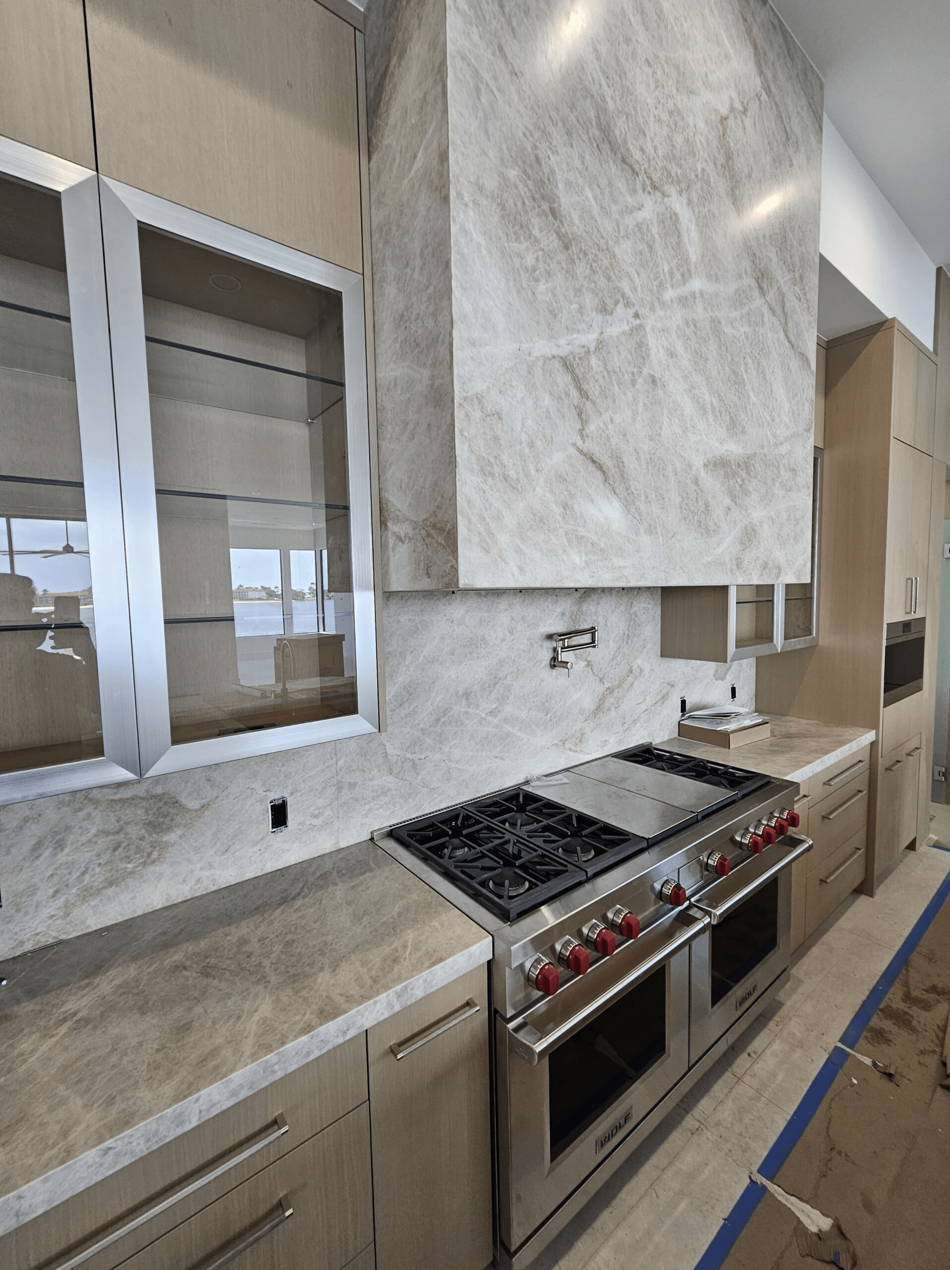 A white marble accented kitchen space with a new stainless steel double range and oven. Above the range is a large marble hood and the white cabinets have glass doors.