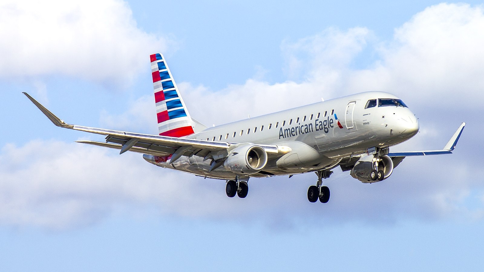 An American Eagle Embraer E175 on final approach with landing gear deployed, framed against a partly cloudy sky.