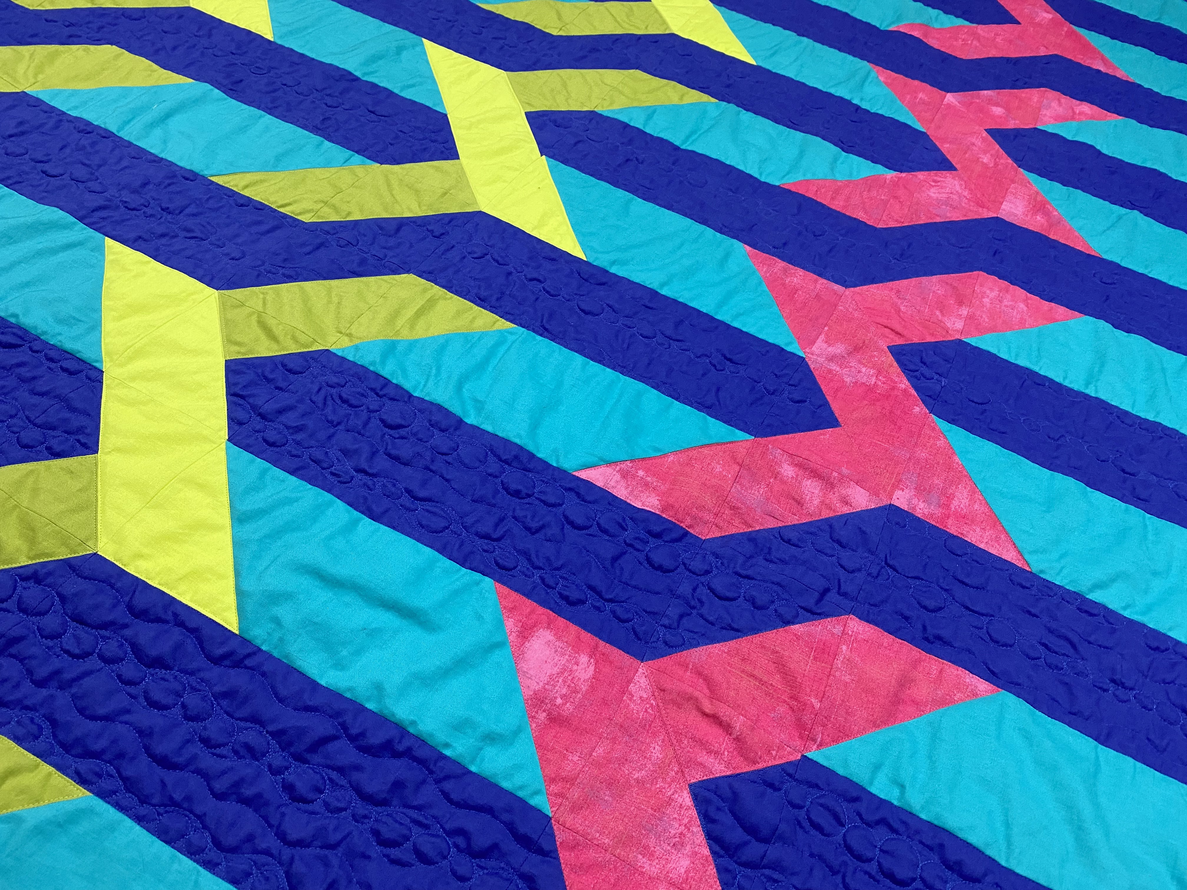 A closeup detail photograph of a quilt in greens, blues, and pink with details of the wave and bubble quilting.