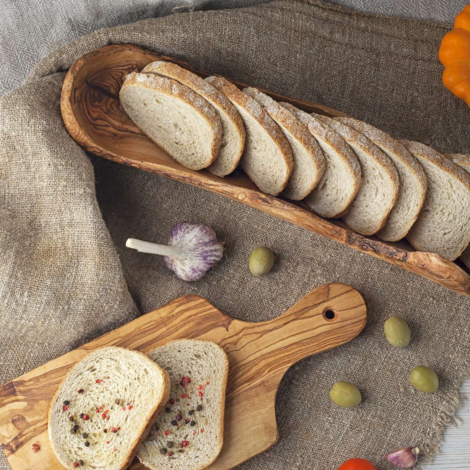 Sliced bread in a wooden bowl beside a cutting board with a sandwich, surrounded by garlic, olives, and vegetables on a cloth.