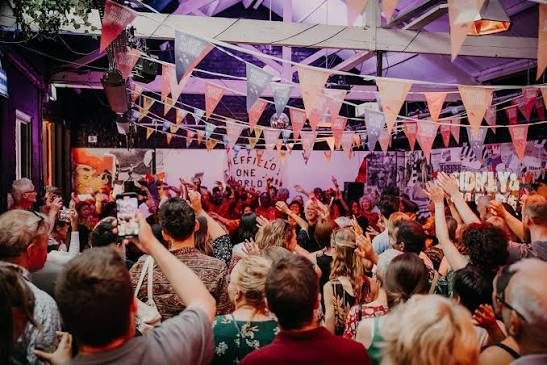 Migration Matters Festival, an audience of people pictured cheering in the foreground, orange bunting overhead, with performers who appear to be dancing on a stage in the background.