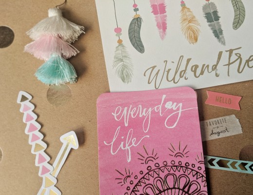 A collection of colorful stationery items on a brown surface. Includes a pink notebook labeled "everyday life," cards with feather designs, an August-themed white tassel decoration with pastel ends, arrow-shaped paper strips, and small banners with words like "HELLO" and "favourite things.